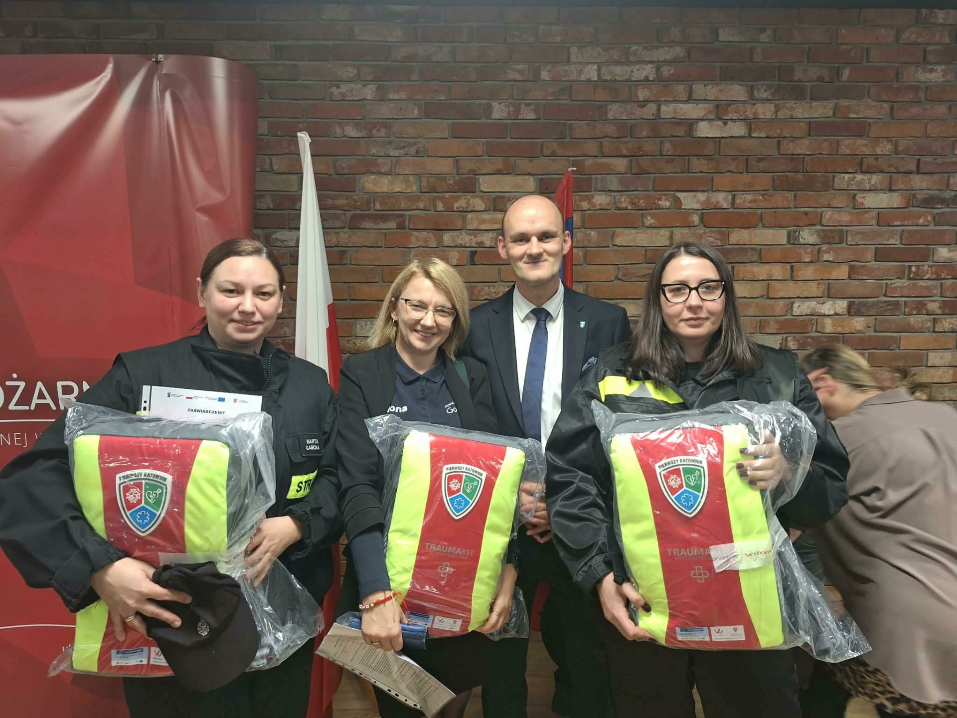 Zdjęcie przedstawia druhny i druhów z plecakami Pierwszego Ratownika i osoby je wręczające
