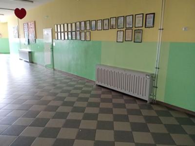 Szkolny korytarz z zielono-żółtymi ścianami, kafelkową podłogą, tablicą z dyplomami i widoczną instalacją grzewczą