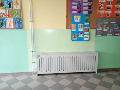 Korytarz szkolny z zielono-żółtymi ścianami, tablicami z pracami uczniów oraz osłoniętym kaloryferem. Widoczne elementy instalacji grzewczej.