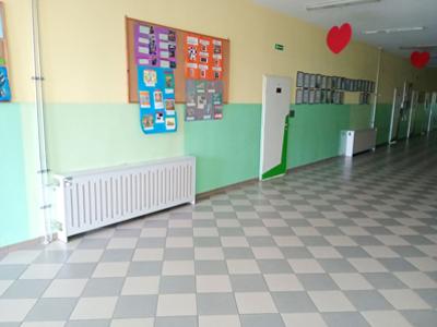 Korytarz szkolny z zielono-żółtymi ścianami, tablicami z pracami uczniów oraz osłoniętym kaloryferem. Widoczne elementy instalacji grzewczej.