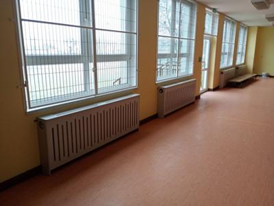 Szkolny korytarz z dużymi oknami w kratach, osłoniętymi kaloryferami, żółtymi ścianami i pomarańczową podłogą