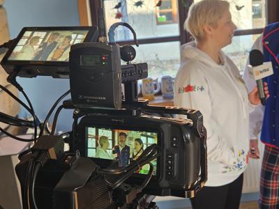 Kobieta w białej bluzie z haftem kociewskim udziela wywiadu dla TV, w kadrze kamera z podglądem rozmowy i osoby w strojach kociewskich