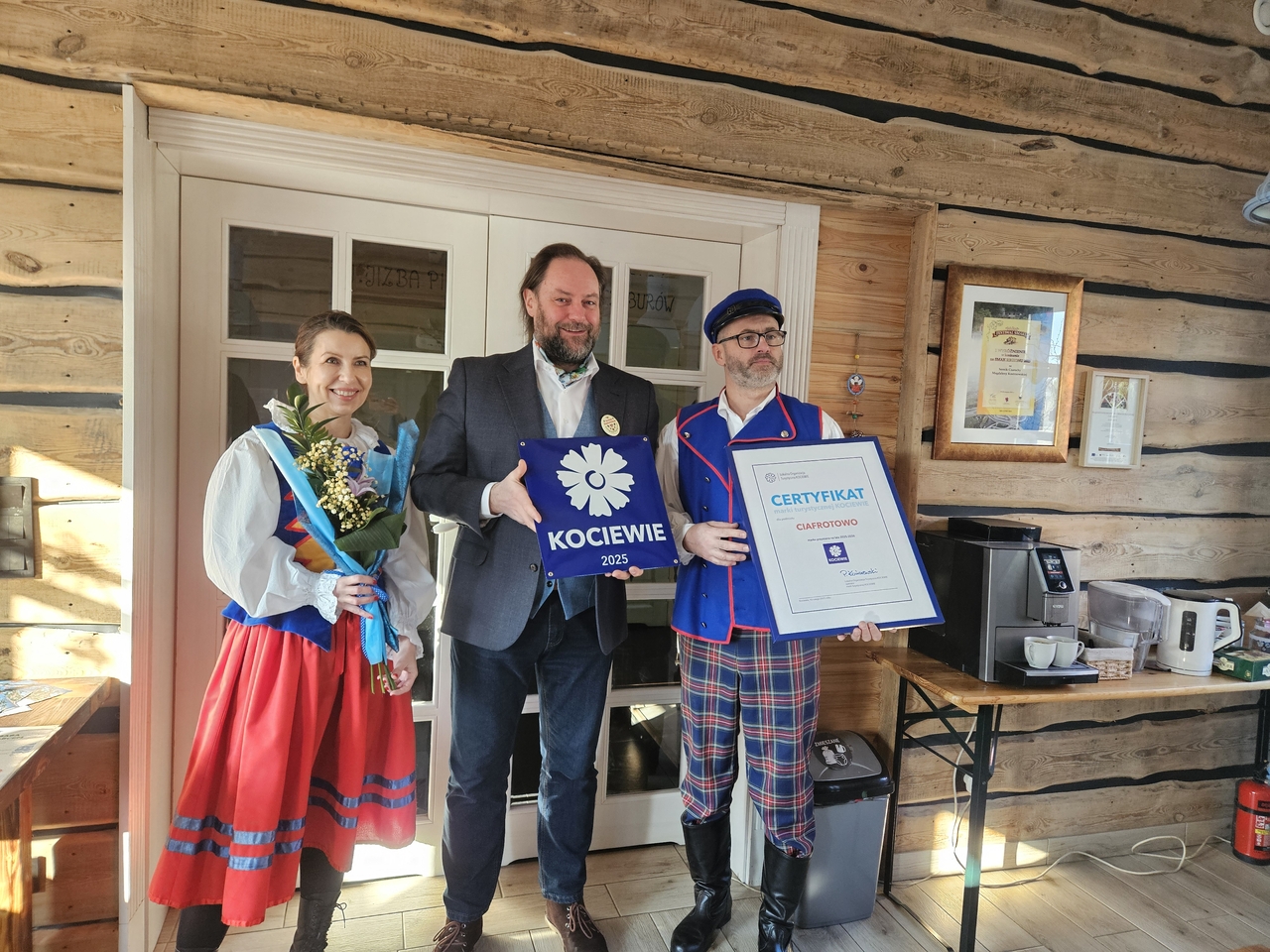 Trzy osoby w drewnianym wnętrzu, dwie w strojach kociewskich. Trzymają tabliczkę „KOCIEWIE 2025”, certyfikat i bukiet kwiatów