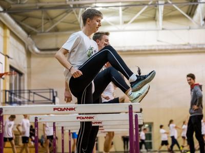 Dwóch młodych sportowców skacze przez płotki w hali sportowej podczas treningu lekkoatletycznego, w tle trener i inni uczestnicy