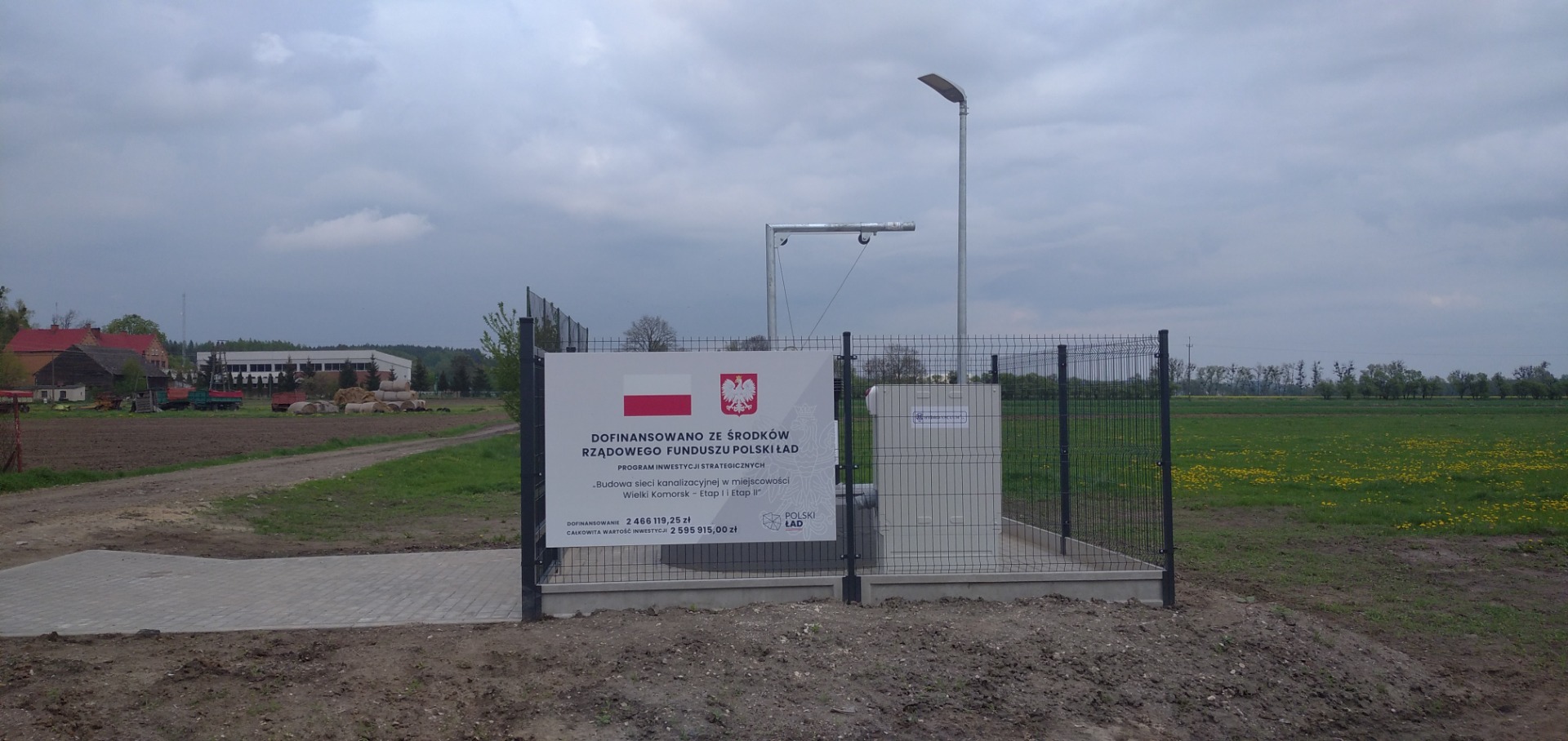 Zdjęcie przedstawia ogrodzenie przepompowni wraz z tablicą z napisem