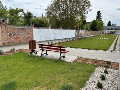 Zdjęcie przedstawiia fragment parkingu, widoczna jest trawa, nasadzone rośliny oraz ławka