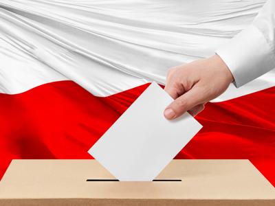 Zdjęcie przedstawia dłoń z kartą do głosowania wkładaną do urny wyborczej, na tle flagi Polski
