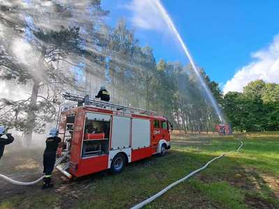 Ćwiczenia OSP, wóz strażacki na tle drzew w obszarze leśnym, z wozu strażackiego puszczana jest woda