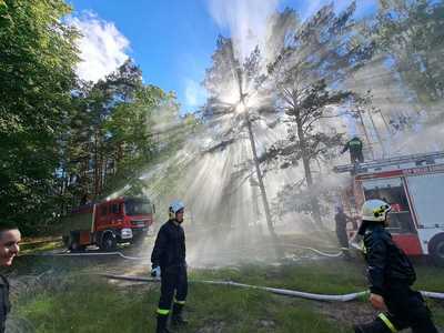 Ćwiczenia OSP, dwa wozu strażackie na tle drzew w obszarze leśnym, z wozów strażackich puszczana jest woda, na pierwszym planie dwóch strażaków OSP