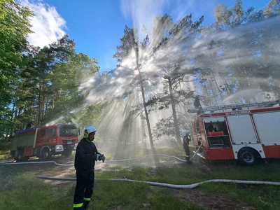 Ćwiczenia OSP, dwa wozu strażackie na tle drzew w obszarze leśnym, z wozów strażackich puszczana jest woda, na pierwszym planie strażak OSP