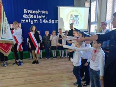 Dzień Edukacji Narodowej w SP Lipinki. Dzieci wyciągają ręce w kierunku sztandaru szkoły, w tle dekoracja i napis Brzechwa dzieciom małym i dużym