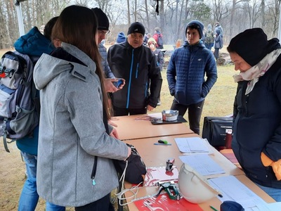 Grupa uczestników przy stoliku organizatorów marszu na orientację, sprawdzających wyniki i sprzęt w terenie leśnym.