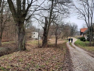 Dwoje uczestników marszu na orientację idzie brukowaną drogą obok starych budynków i lasu w kierunku opuszczonego budynku
