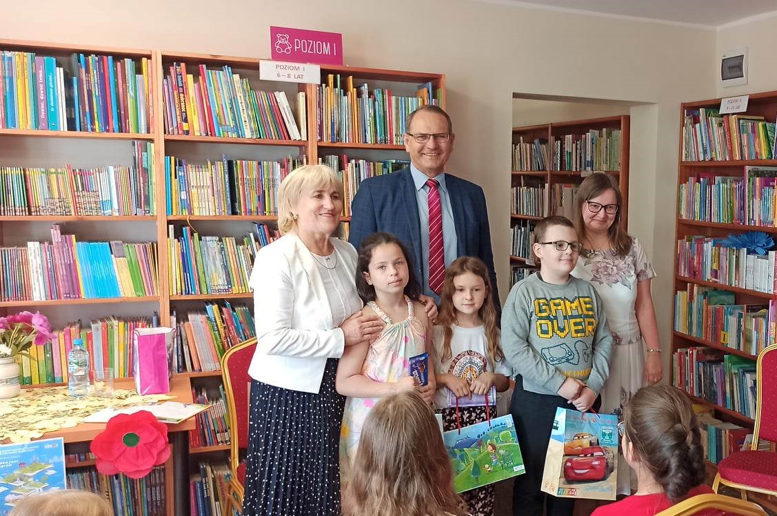 podsumowanie konkursu "Pożeracze książek", wójt gminy, przewodnicząca gminy, dzieci w bibliotece na tle regałów z książkami