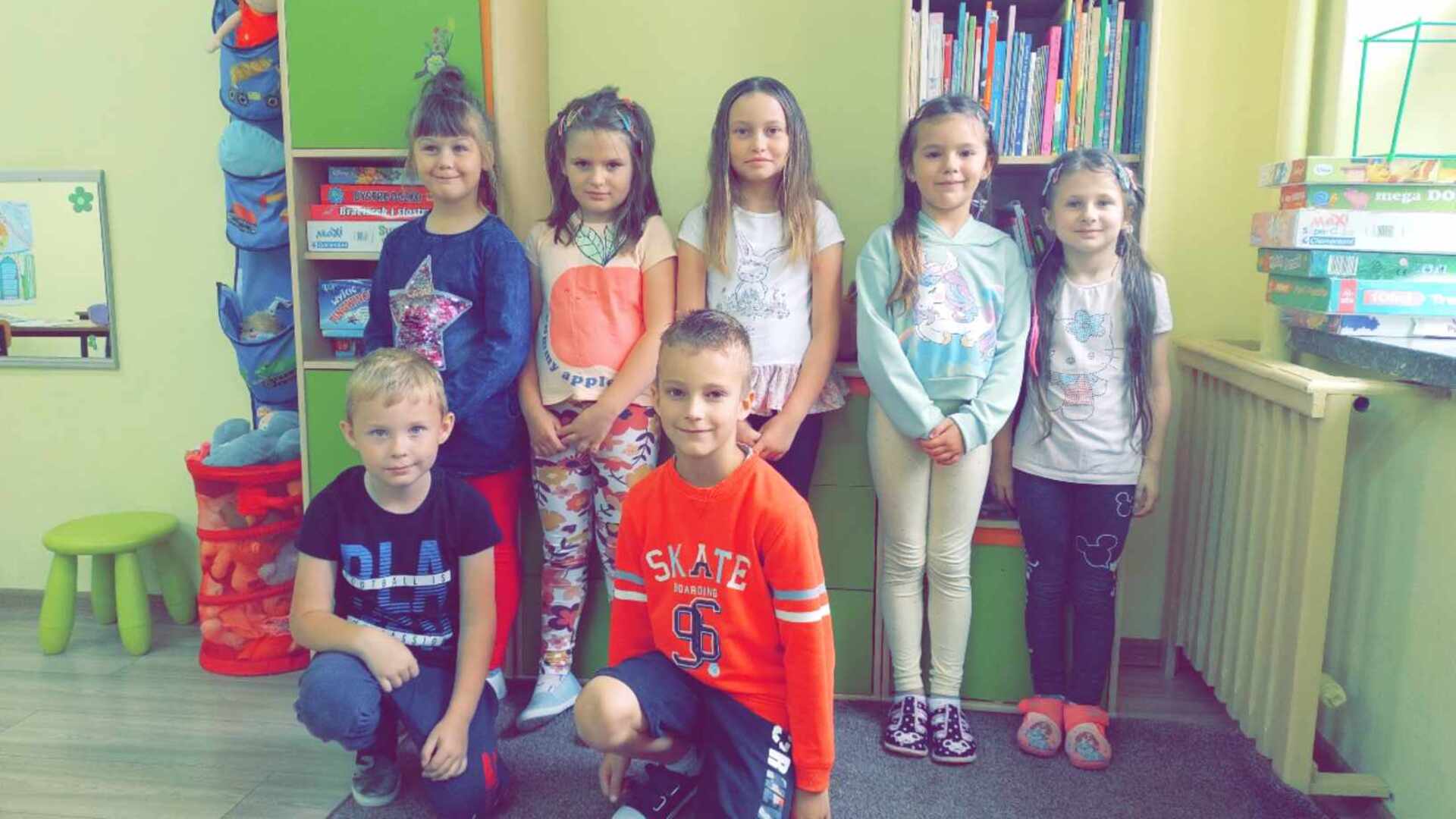 Dzień Fryzjera. Grupa uśmiechniętych dzieci pozuje w bibliotece lub świetlicy, otoczona książkami i grami planszowymi