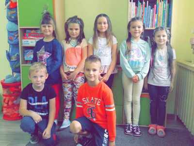 Dzień Fryzjera. Grupa uśmiechniętych dzieci pozuje w bibliotece lub świetlicy, otoczona książkami i grami planszowymi