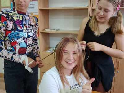 Dzień Fryzjera. Dwie dziewczyny zaplatają warkocze koleżance, która uśmiecha się i pokazuje kciuk w górę w geście aprobaty
