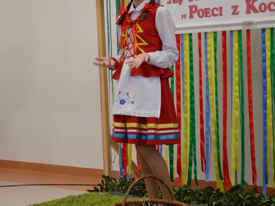 Dziewczyna w stroju ludowym z wiankiem na głowie recytuje na scenie konkursowej, gestykulując, przed dekoracją z kwiatami i wstążkami