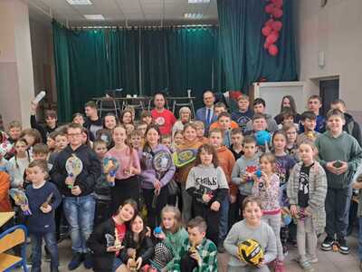 Turniej szachowy. Grupa dzieci i dorosłych na sali gimnastycznej, trzymających nagrody i sportowe akcesoria, udekorowana scena w tle.