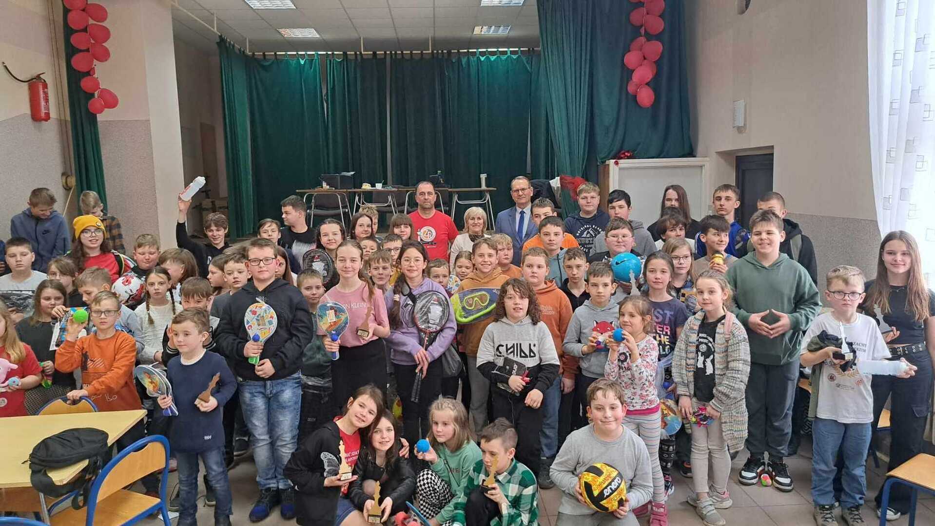 Turniej szachowy. Grupa dzieci i dorosłych na sali gimnastycznej, trzymających nagrody i sportowe akcesoria, udekorowana scena w tle.