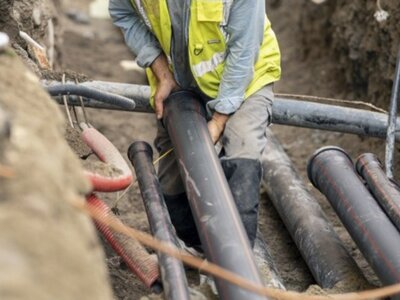 Pracownik budowlany w żółtej kamizelce układa rury w wykopie, wokół inne przewody i elementy infrastruktury podziemnej