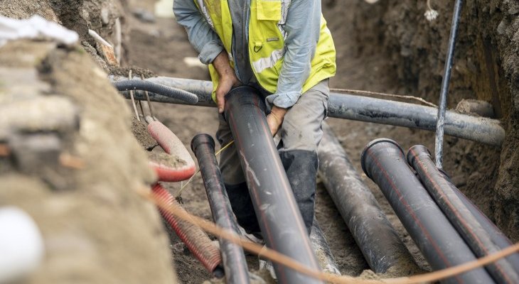 Pracownik budowlany w żółtej kamizelce układa rury w wykopie, wokół inne przewody i elementy infrastruktury podziemnej