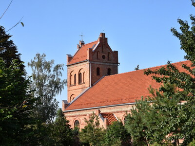 Kościół w Warlubiu. Ceglany kościół z wieżą i dachem, otoczony drzewami, na tle błękitnego nieba.