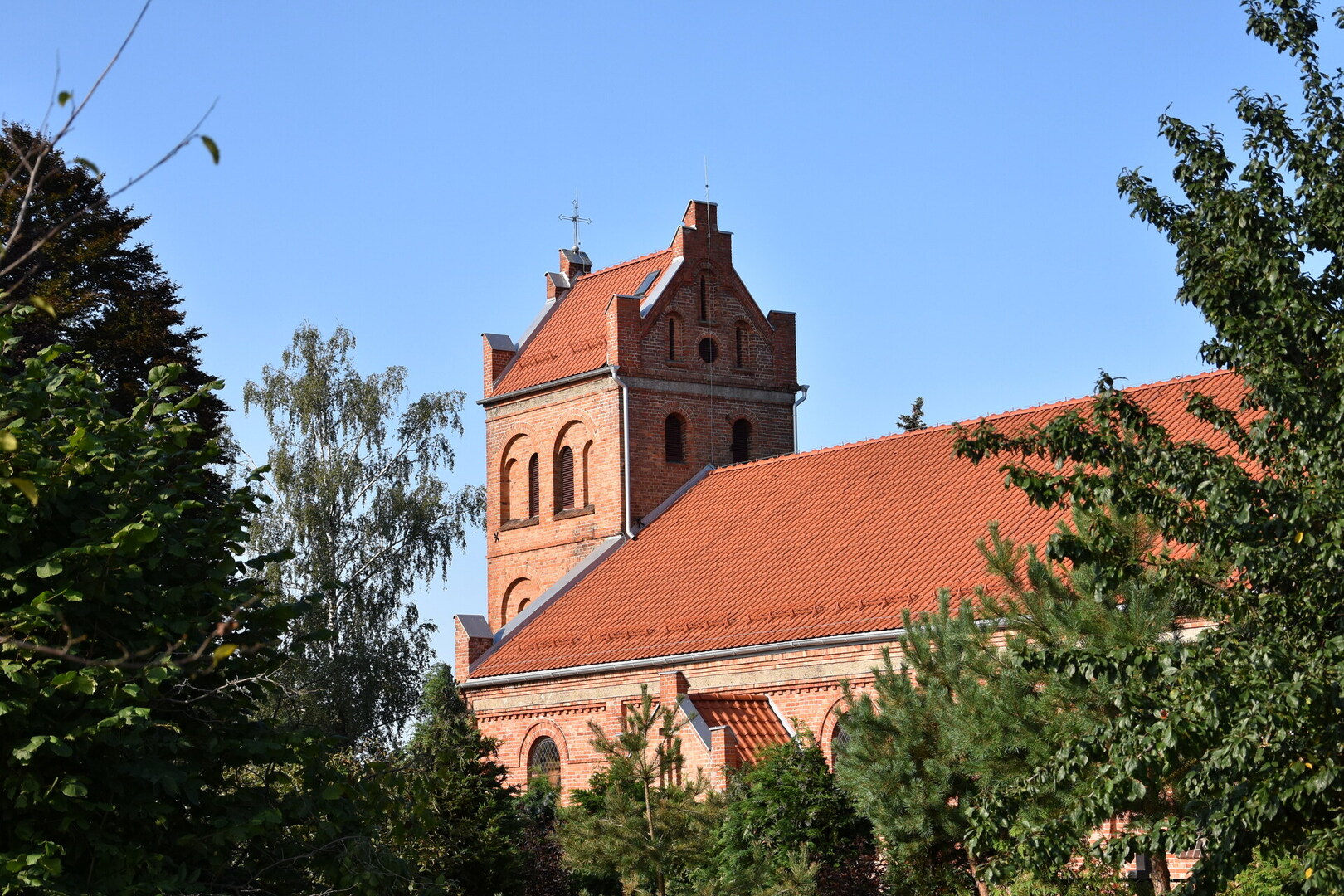Kościół w Warlubiu. Ceglany kościół z wieżą i dachem, otoczony drzewami, na tle błękitnego nieba.