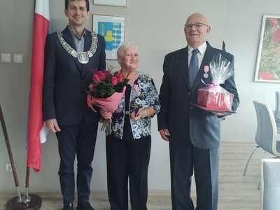 Wójt Gminy z uśmiechniętą parą podczas wręczenia medali. Kobieta trzyma bukiet róż, mężczyzna medal i prezent. W tle flaga Polski i herb gminy
