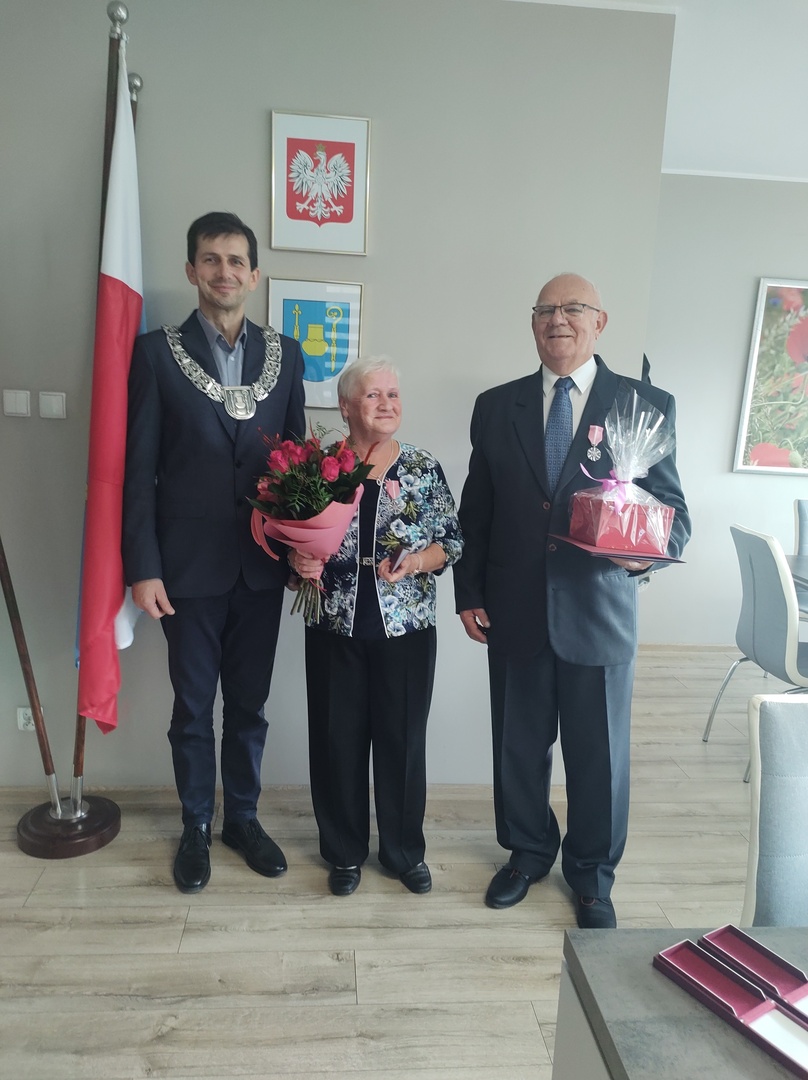 Wójt Gminy z uśmiechniętą parą podczas wręczenia medali. Kobieta trzyma bukiet róż, mężczyzna medal i prezent. W tle flaga Polski i herb gminy