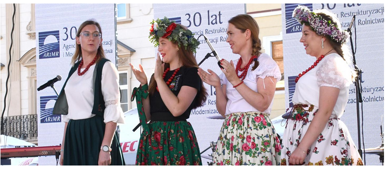 Cztery kobiety w strojach ludowych na scenie w tle baner i mikrofony