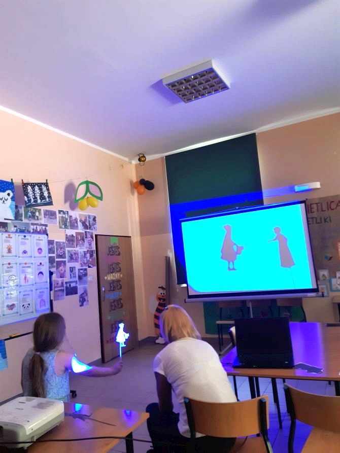 Zdjęcie przedstawia dziecko i wychowawczynię obserwujące teatr cieni na tablicy interaktywnej.