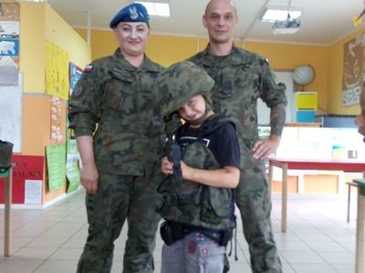 Grafika przedstawia dwóch żołnierzy i wychowanka Placówki Wsparcia Dziennego w Laskach przebranego za żołnierza. 