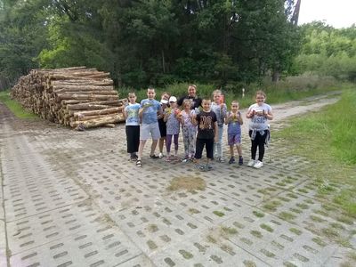 Zdjęcie przedstawia wychowanków Placówki Wsparcia Dziennego z Lasek wraz z wychowawczynią podczas zajęć teatralno - dziennikarskich w leśnej scenerii.