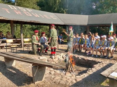 Zdjęcie przedstawia dwójkę harcerzy składających przyrzeczenie oraz innych harcerzy biorących udział w spotkaniu integracyjnym przy wiatce na boisku w Ostrowcu. 
