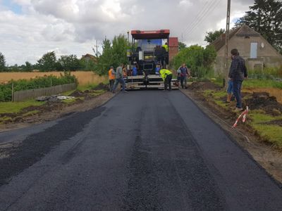 Zdjęcie przedstawia remontowany odcinek drogi w Malechówku, osoby zaangażowane w pracę oraz specjalistyczną maszynę od utwardzania nawierzchni.