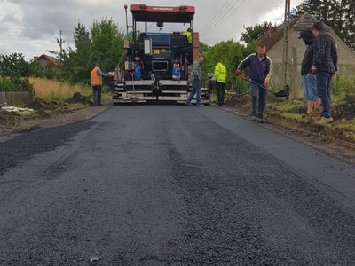 Zdjęcie przedstawia remontowany odcinek drogi w Malechówku, osoby zaangażowane w pracę oraz specjalistyczną maszynę od utwardzania nawierzchni. 