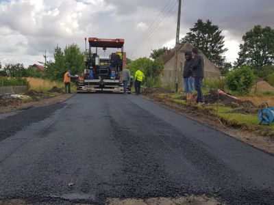 Zdjęcie przedstawia remontowany odcinek drogi w Malechówku, osoby zaangażowane w pracę oraz specjalistyczną maszynę od utwardzania nawierzchni. 