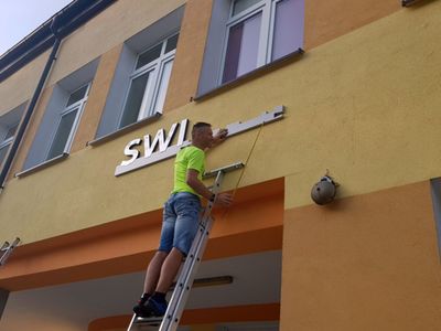 Zdjęcie przedstawia montaż napisu Świetlica Kultury w Malechowie na początkowym etapie oraz mężczyznę na drabinie. 