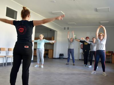 Zdjęcie przedstawia ćwiczenia ruchowe seniorek z Malechowa, które gimnastykują się pod okiem fizjoterapeuty. 