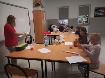 Zdjęcie przedstawia prowadzącą i grupę dzieci podczas nauki nazw owoców z j. angielskiego.