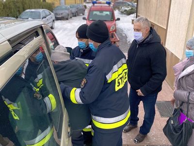 Strażacy z OSP Malechowo pomagają seniorowi, który przyjechał zaszczepić się przeciw COVID-19 podczas wysiadania z auta.
