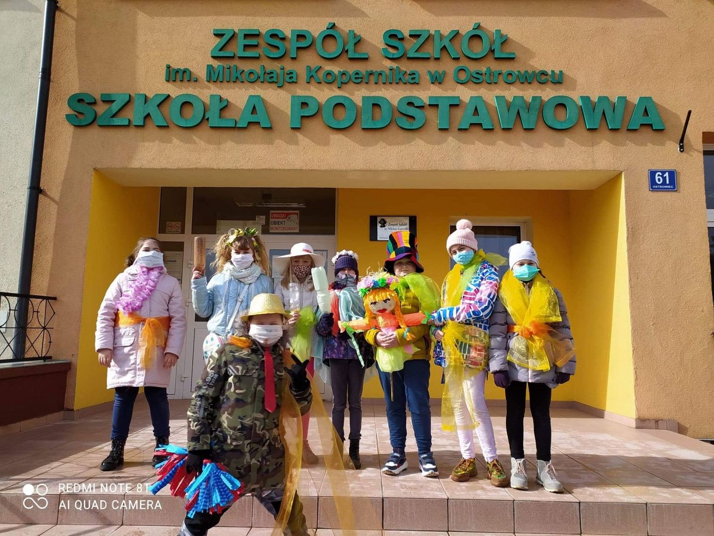 Grupa uczniów z Zespołu Szkolnego w Ostrowcu przed budynkiem szkoły gotowa do powitania wiosny. 