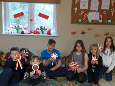  Dzieci z Placówki Wsparcia Dziennego w Kosierzewie z kotylionami, a w tle gazetka z okazji majowych świąt i udekorowane okno.