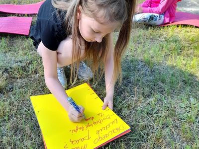 Dziewczynka z Placówki Wsparcia Dziennego w Sulechowie pisząca na kartce, w tle dwie dziewczynki siedzące na matach gimnastycznych.
