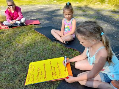 Trzy dziewczynki z Placówki Wsparcia Dziennego w Sulechowie, siedzące na matach gimnastycznych na zewnątrz placówki w Sulechowie. Na pierwszym planie dziewczynka uzupełnia plakat.