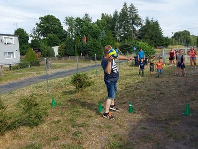 Tatusiowe biorący udział w sportowych konkurencjach na wesoło przed świetlicą, w tle dzieci. 