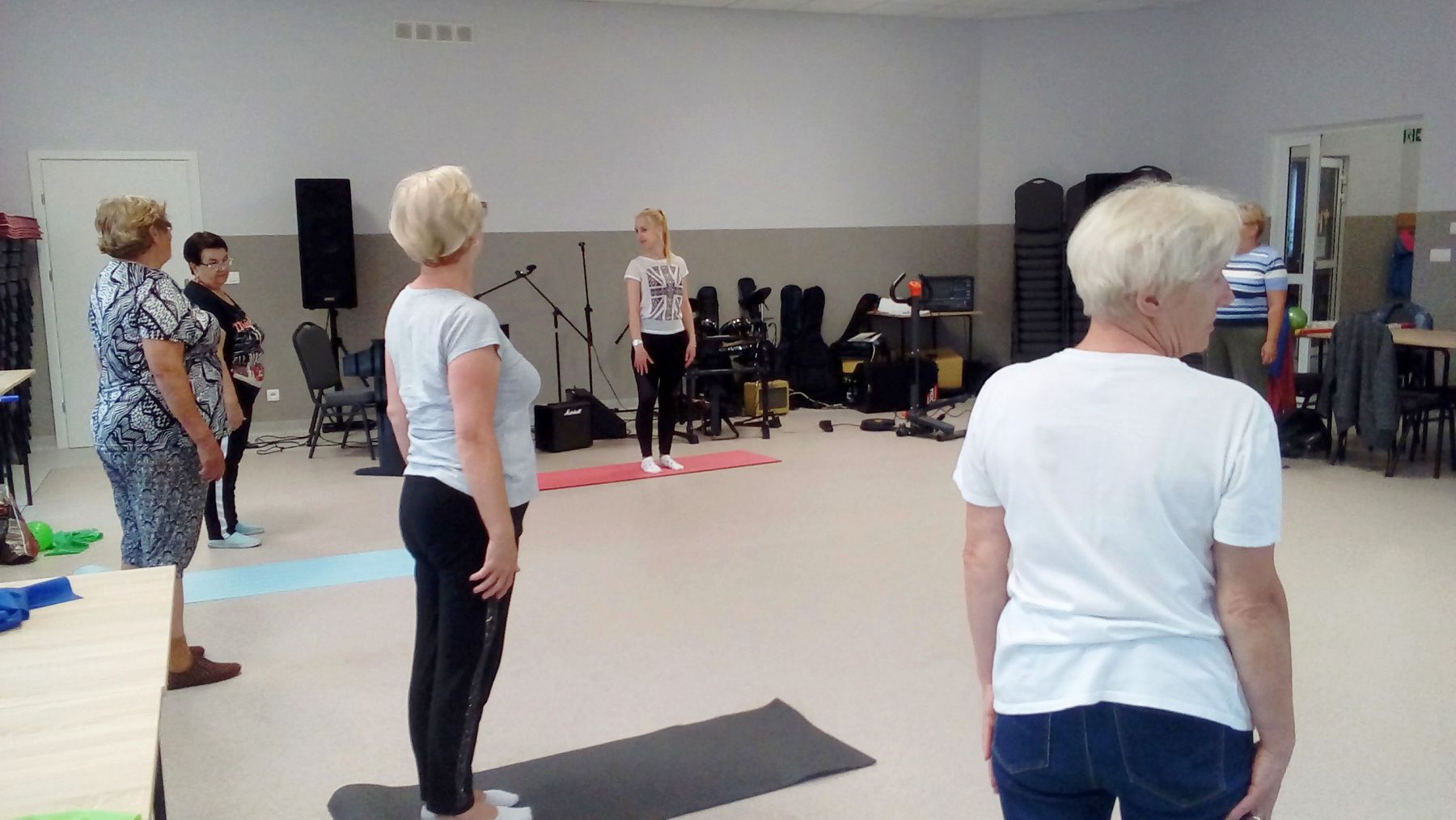 Seniorzy podczas ćwiczeń gimnastycznych, w tle fizjoterapeutka prowadząca zajęcia.