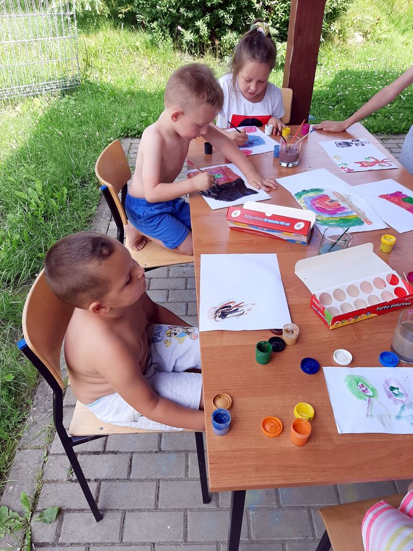 Dzieci z Placówki Wsparcia Dziennego w Przystawach malują farbami przy stoliku na tarasie świetlicy wiejskiej w Przystawach. 