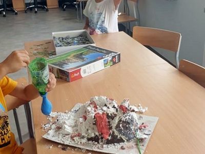 Chłopiec pozuje z balonem, na pierwszym planie wykonany z pianki do golenia wulkan na ławce szkolnej. 